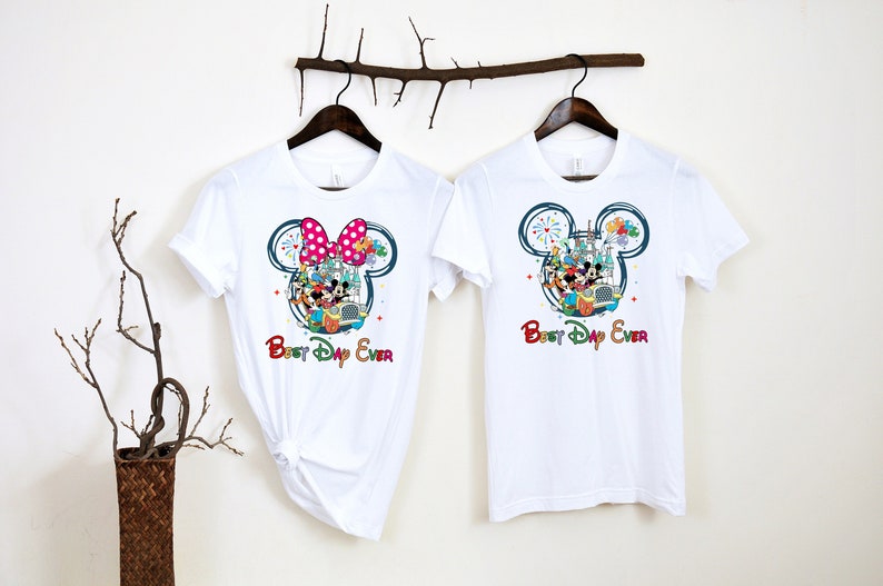 May include: Two white t-shirts with a Mickey Mouse and Minnie Mouse design. The shirts feature a colorful illustration of Mickey and Minnie Mouse with other Disney characters in a car. The text "Best Day Ever" is printed on both shirts.