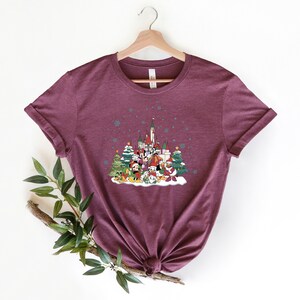 May include: A maroon t-shirt with a Christmas-themed design featuring Mickey Mouse and friends in front of a castle. The design includes a snowy scene with Christmas trees and snowflakes.