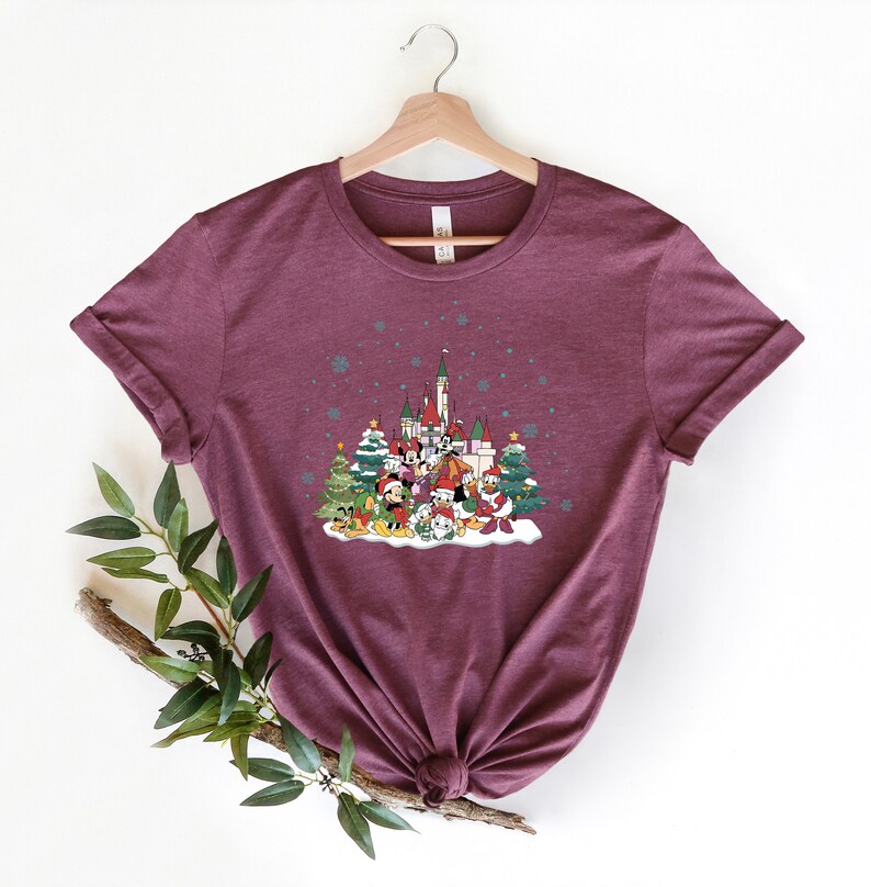 May include: A maroon t-shirt with a Christmas-themed design featuring Mickey Mouse and friends in front of a castle. The design includes a snowy scene with Christmas trees and snowflakes.