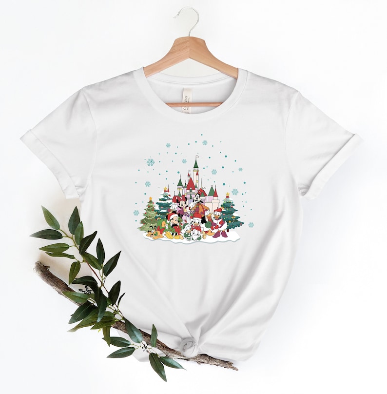 May include: White t-shirt with a Christmas-themed graphic featuring Mickey Mouse and friends in front of a snow-covered castle. The graphic includes a Christmas tree, presents, and snowflakes.