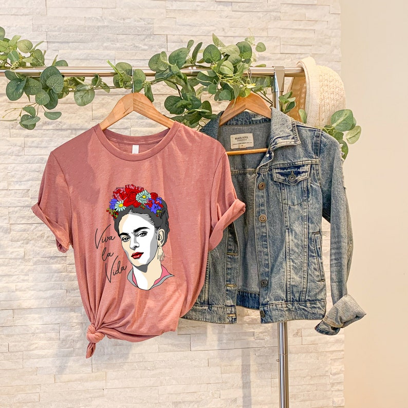 May include: A dusty rose t-shirt with a graphic of Frida Kahlo wearing a flower crown and the text "Viva la Vida".