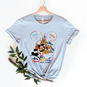 Puede incluir: Camiseta azul claro con un gr&aacute;fico de Mickey Mouse y sus amigos frente al Castillo de Cenicienta. El texto "2024" y "NAME" est&aacute;n en las esquinas superiores del gr&aacute;fico. El texto "Disney Family Vacation" est&aacute; debajo del gr&aacute;fico.