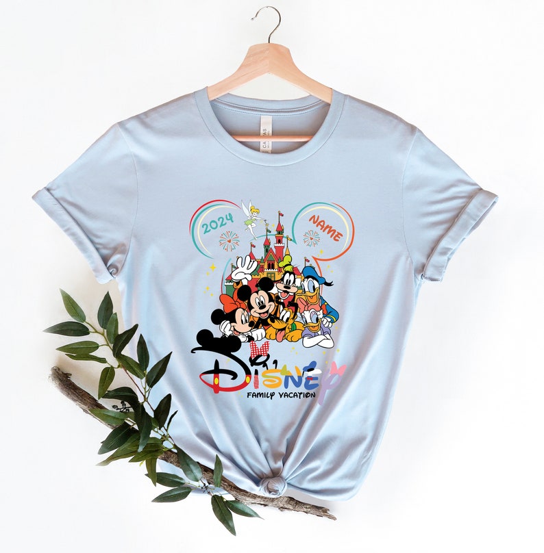Puede incluir: Camiseta azul claro con un gr&aacute;fico de Mickey Mouse y sus amigos frente al Castillo de Cenicienta. El texto "2024" y "NAME" est&aacute;n en las esquinas superiores del gr&aacute;fico. El texto "Disney Family Vacation" est&aacute; debajo del gr&aacute;fico.