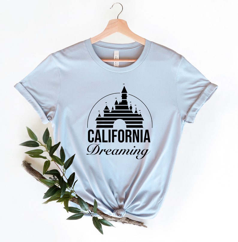 Puede incluir: Camiseta azul claro con una silueta negra del Castillo de la Bella Durmiente y el texto "CALIFORNIA Dreaming".