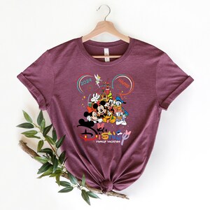 Puede incluir: Una camiseta de color marr&oacute;n rojizo con un gr&aacute;fico de Mickey Mouse, Minnie Mouse, Donald Duck, Goofy y Pluto frente al Castillo de Cenicienta. El texto "Disney Family Vacation" est&aacute; debajo de los personajes. Los textos "2024" y "Nombre" est&aacute;n en c&iacute;rculos encima de los personajes.