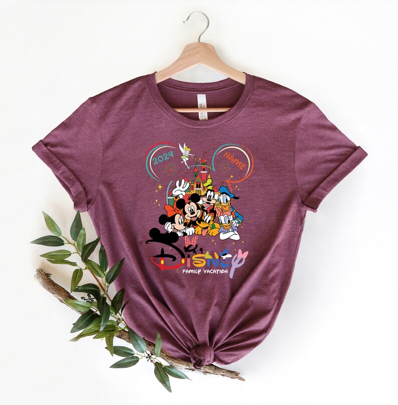 Puede incluir: Una camiseta de color marr&oacute;n rojizo con un gr&aacute;fico de Mickey Mouse, Minnie Mouse, Donald Duck, Goofy y Pluto frente al Castillo de Cenicienta. El texto "Disney Family Vacation" est&aacute; debajo de los personajes. Los textos "2024" y "Nombre" est&aacute;n en c&iacute;rculos encima de los personajes.