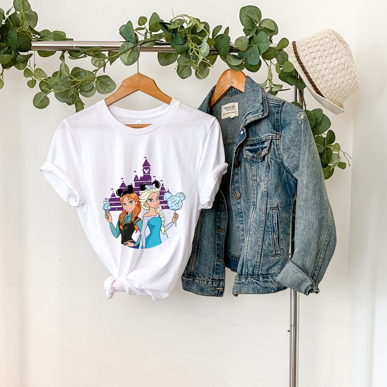 以下が含まれることがあります： ミッキーマウスの耳をつけ、ミッキーマウスの形をしたアイスクリームを持っているアナとエルサのフロストンのグラフィックが描かれた白いTシャツ。グラフィックには、シンデレラ城の紫色のシルエットも含まれています。