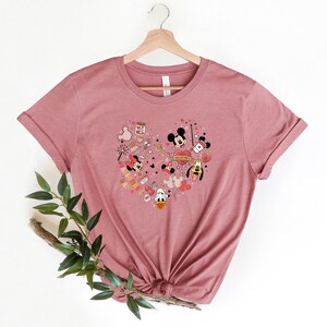Pode incluir: Uma camiseta rosa com um design em forma de cora&ccedil;&atilde;o que apresenta Mickey Mouse, Minnie Mouse e Pateta. O design inclui v&aacute;rios elementos tem&aacute;ticos do Dia dos Namorados, como cora&ccedil;&otilde;es, bal&otilde;es e uma chave. O texto "Happy Valentine's Day" est&aacute; inclu&iacute;do no design.