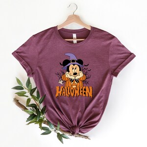 K&ouml;nnte beinhalten: Ein maroonfarbenes T-Shirt mit einem Aufdruck von Mickey Mouse als Hexe mit einem K&uuml;rbis und dem Text "HALLOWEEN".