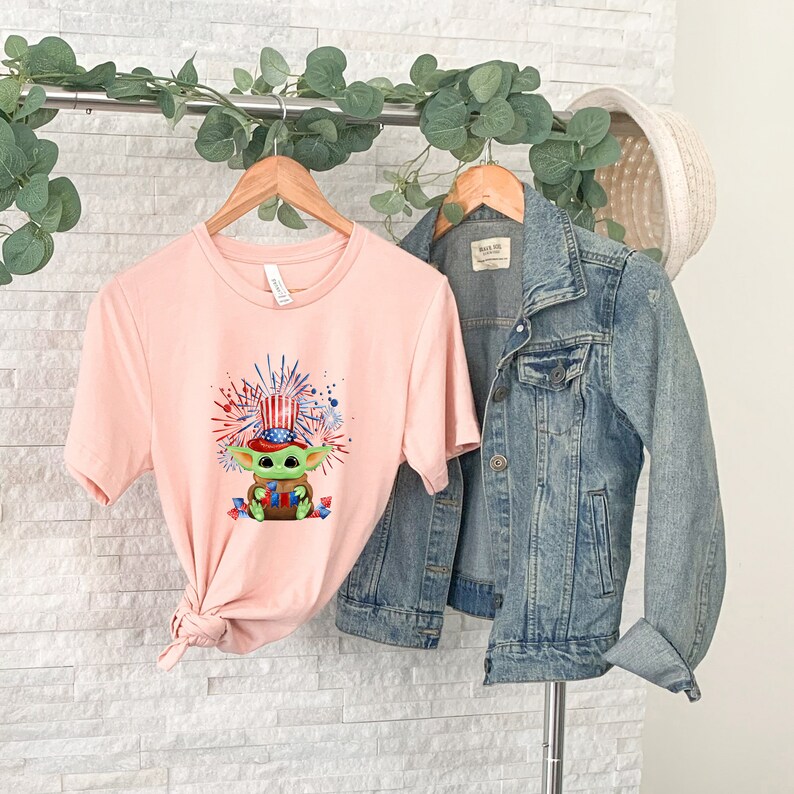 May include: A pink t-shirt with a cartoon image of a green character wearing a red, white, and blue hat with fireworks in the background. The shirt is hanging on a wooden hanger with a denim jacket hanging next to it.