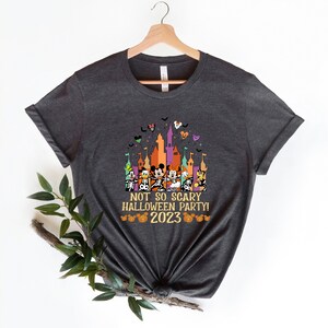 May include: A dark grey t-shirt featuring a colourful graphic of Mickey Mouse and his friends in front of a castle. The text "Not so scary Halloween party! 2023" is printed below the graphic.