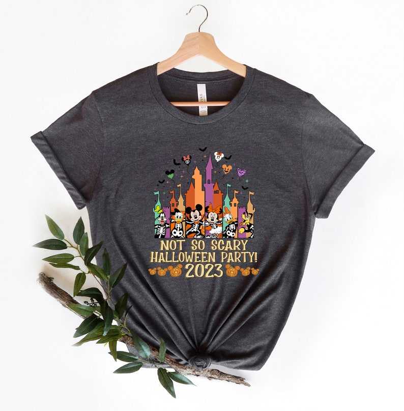 May include: A dark grey t-shirt featuring a colourful graphic of Mickey Mouse and his friends in front of a castle. The text "Not so scary Halloween party! 2023" is printed below the graphic.