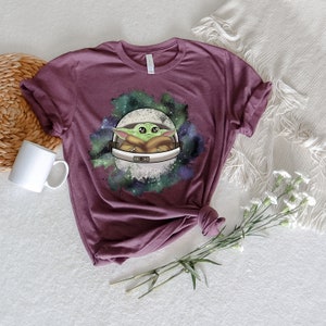 May include: A maroon t-shirt with a graphic of a green alien character in a space suit, surrounded by a galaxy-like design with stars and nebulae.