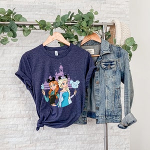 以下が含まれることがあります： シンデレラ城の前でディズニーのプリンセス、アナとエルサが描かれたネイビーブルーのTシャツ。プリンセスはミッキーマウスの耳と綿菓子を持っています。
