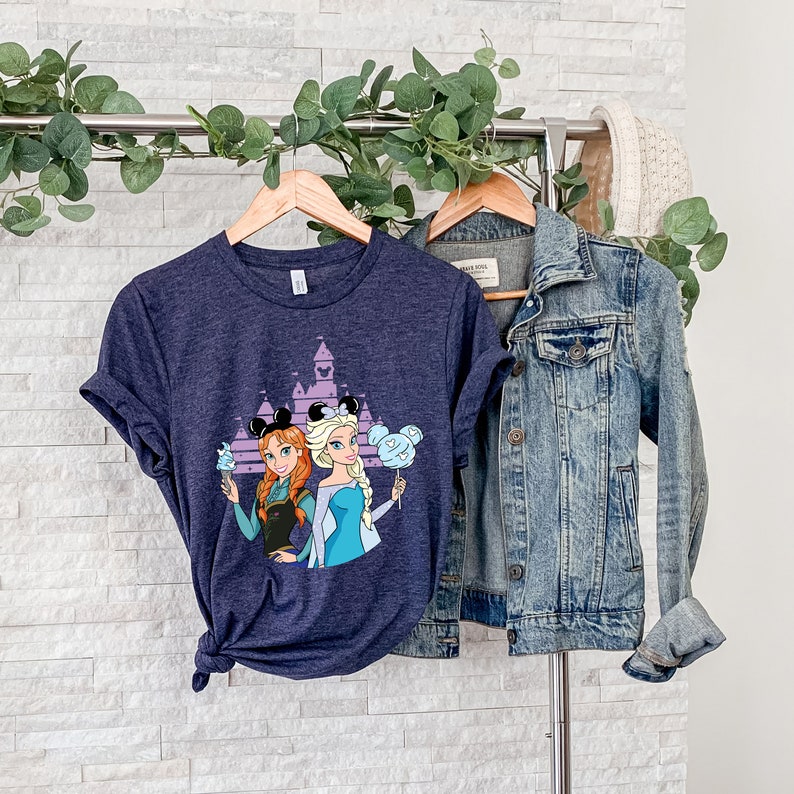 以下が含まれることがあります： シンデレラ城の前でディズニーのプリンセス、アナとエルサが描かれたネイビーブルーのTシャツ。プリンセスはミッキーマウスの耳と綿菓子を持っています。
