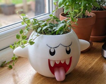 Puede incluir: Una maceta blanca y esférica con forma de personaje fantasma, con la boca abierta, dientes afilados, lengua rosa y ojos enfadados. La maceta contiene una planta verde colgante y está situada junto a otras hierbas en macetas y una cafetera.