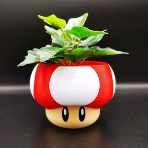 May include: A red and white mushroom-shaped planter with a light brown base, holding a green leafy plant. The planter has two white circles on the sides and black oval eyes. A decorative item for home decor.