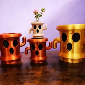 Puede incluir: Tres macetas impresas en 3D con forma de personaje con una expresión de sorpresa. Las macetas son de diferentes colores: dorado, cobre y blanco. La maceta blanca tiene una pequeña planta que crece en ella.