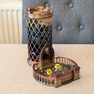 May include: A 3D printed dice tower with a black and gold colour scheme. The tower is shaped like a castle with a spiral staircase and a small balcony. Two yellow dice are on the balcony.