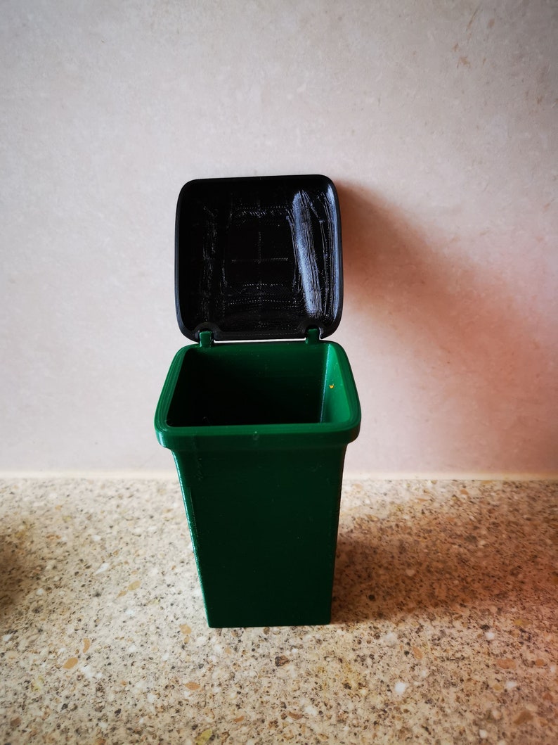Op de afbeelding: Een groene prullenbak met een zwarte deksel. De deksel is open en de prullenbak is leeg.