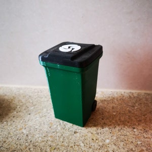 Op de afbeelding: Een kleine groene recyclebak met een zwart deksel en een wit recycle-symbool. De bak staat op een gevlekt aanrecht.