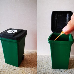 Op de afbeelding: Een kleine groene prullenbak met een zwart deksel en een wit recycle-symbool. Het deksel is open en een klein oranje stokje wordt erin geplaatst.