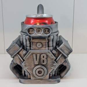 May include: A silver-colored V8 engine-shaped can cooler holding a red soda can. The cooler has detailed engine components and the text "V8" on the front. The design is a unique novelty item.