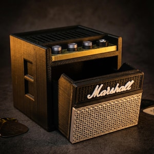 Op de afbeelding: Een miniatuur zwarte en gouden Marshall-versterker replica met de merknaam in witte letters. De versterker heeft zilveren knoppen en een luidsprekerroosterontwerp. Een plectrum ligt in de buurt.