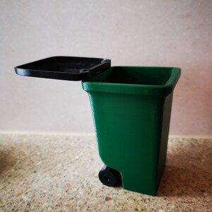 Op de afbeelding: Een kleine groene prullenbak met een zwart deksel en een zwart wiel. Het deksel is open en de prullenbak staat op een gevlekt aanrecht.