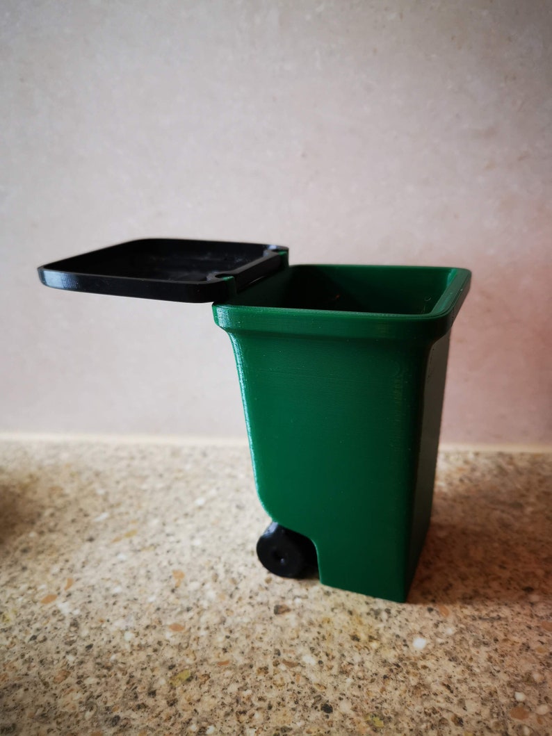 Op de afbeelding: Een kleine groene prullenbak met een zwart deksel en een zwart wiel. Het deksel is open en de prullenbak staat op een gevlekt aanrecht.