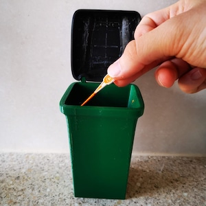 Op de afbeelding: Een groene plastic prullenbak met een zwart deksel. Het deksel is open en een klein oranje en wit voorwerp wordt in de prullenbak gegooid.