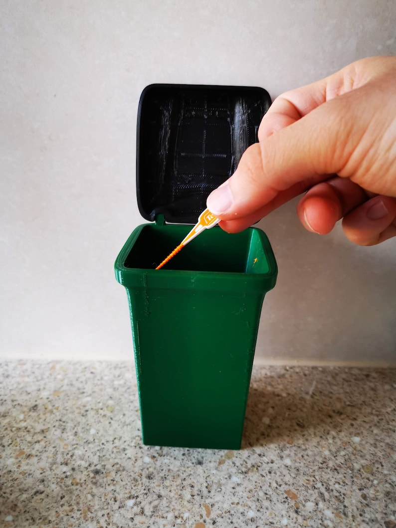 Op de afbeelding: Een groene plastic prullenbak met een zwart deksel. Het deksel is open en een klein oranje en wit voorwerp wordt in de prullenbak gegooid.