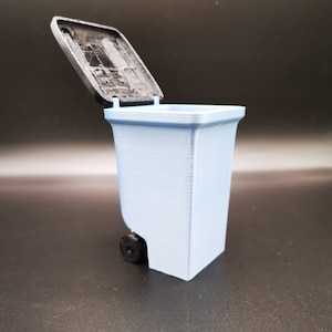 Op de afbeelding: Een lichtblauwe plastic prullenbak met een zwarte deksel. De deksel is open en de prullenbak staat op een zwart oppervlak.