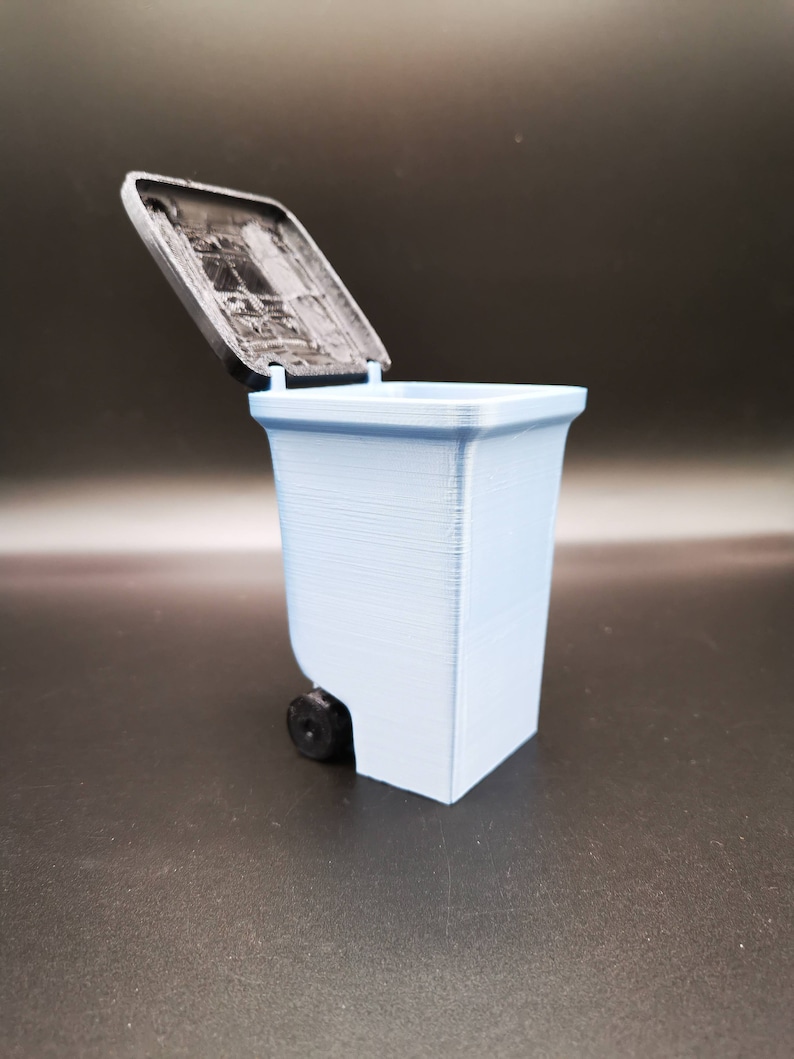 Op de afbeelding: Een lichtblauwe plastic prullenbak met een zwarte deksel. De deksel is open en de prullenbak staat op een zwart oppervlak.