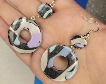 May include: A pair of silver dangle earrings with a black, white, and blue abstract pattern. The earrings feature two circular shapes, one larger than the other, with a hole in the center.