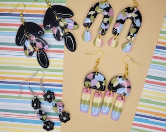 May include: A set of six handmade polymer clay earrings. The earrings are in black, white, pink, and green. The earrings are shaped like arches, flowers, and leaves.