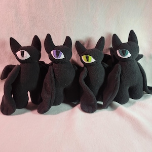 May include: Four black plush bat toys with large, stylized eyes in white, purple, green, and blue. The bats have pointed ears, wings, and small arms and legs. The toys are arranged in a row against a soft, light-colored background.