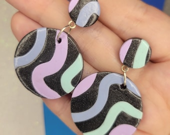May include: A pair of dangle earrings with a black, purple, and blue swirl pattern. The larger earring has a round shape, while the smaller earring is a smaller circle.
