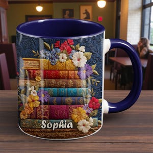 Può includere: Tazza in ceramica con interno e manico blu navy, raffigurante una pila di libri colorati in rilievo adornati da fiori. Il nome "Sophia" è visualizzato sul davanti. La tazza è appoggiata su una superficie di legno.