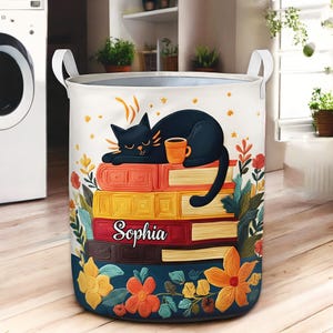 May include: A round laundry hamper with white handles. The hamper features a black cat sleeping on a stack of books, holding a coffee cup. The name "Sophia" is printed on one of the books. The design includes flowers and leaves.