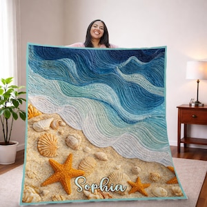 May include: A beach-themed quilt featuring a detailed design of ocean waves, sand, seashells, and starfish. The quilt has a blue and turquoise border and includes the name "Sophia" in cursive script. The overall color palette includes shades of blue, beige, and orange.