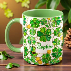 May include: A white ceramic mug with a green handle and rim, decorated with green shamrocks, gold coins, and a leprechaun hat. The name "Sophia" is written in green script on the mug.