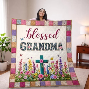 Può includere: Una coperta trapuntata con le parole "Blessed GRANDMA" in corsivo bordeaux e stampa floreale. La coperta presenta una croce, farfalle e un bordo floreale colorato. Lo sfondo è bianco.