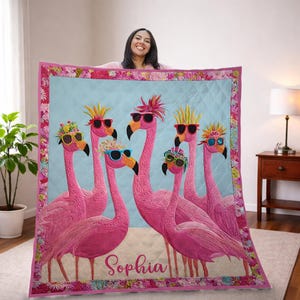 May include: A pink quilt featuring six flamingo illustrations wearing sunglasses and decorative headwear. The word "Sophia" is embroidered on the quilt. The quilt has a light blue center and a floral border.