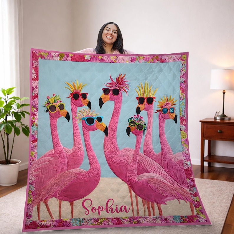May include: A pink quilt featuring six flamingo illustrations wearing sunglasses and decorative headwear. The word "Sophia" is embroidered on the quilt. The quilt has a light blue center and a floral border.
