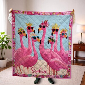 May include: A pink flamingo quilt featuring six flamingos wearing sunglasses and decorative headwear. The quilt has a light blue background with a diamond pattern and a floral border. The name "Sophia" is written at the bottom.
