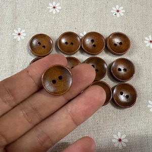 May include: A selection of round, wooden buttons in various sizes. The buttons are a warm brown colour with two holes for sewing. Some buttons are held in a hand, while others are scattered on a light-coloured fabric.