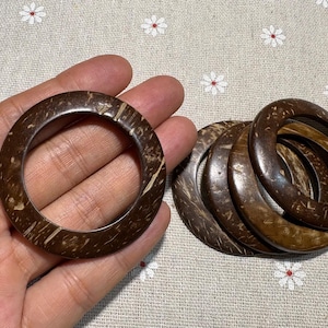 Puede incluir: Varios brazaletes de concha de coco marr&oacute;n oscuro. Uno se sostiene en la mano, los otros est&aacute;n apilados. Los brazaletes tienen un aspecto natural y org&aacute;nico con vetas de madera visibles y miden aproximadamente 7 cm de di&aacute;metro.