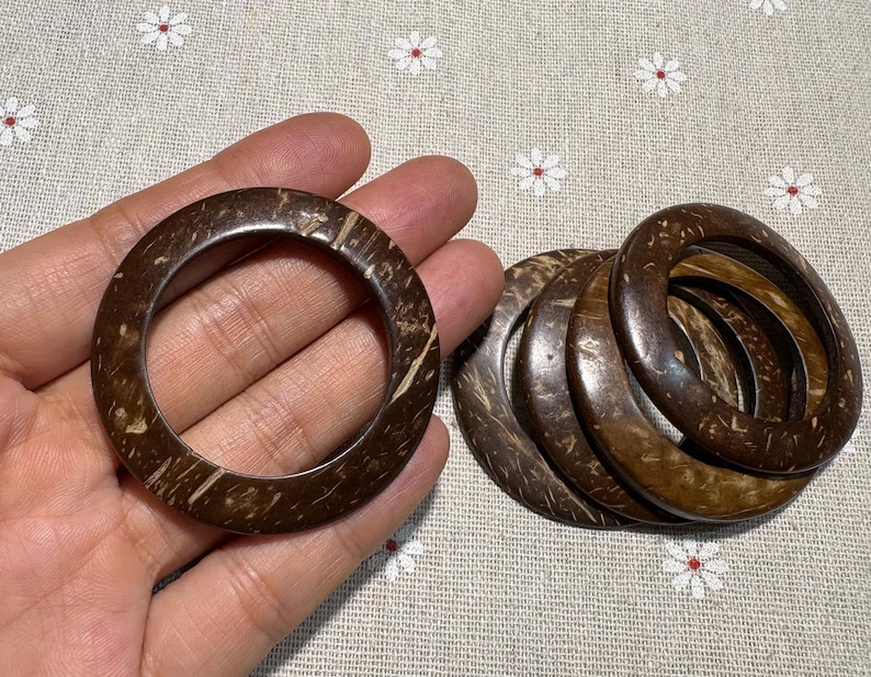 Puede incluir: Varios brazaletes de concha de coco marr&oacute;n oscuro. Uno se sostiene en la mano, los otros est&aacute;n apilados. Los brazaletes tienen un aspecto natural y org&aacute;nico con vetas de madera visibles y miden aproximadamente 7 cm de di&aacute;metro.