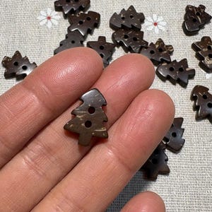 Könnte beinhalten: Eine Sammlung dunkelbrauner, baumförmiger Knöpfe. Die Knöpfe haben drei Löcher und bestehen aus einem natürlichen Material. Mehrere Knöpfe sind auf einem hellen Stoff mit floralen Mustern verstreut.